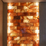 A rectangular wall-mounted light panel made of stacked warm-toned stone tiles glowing with amber, orange, tan, and cream light, set between dark gray side walls.