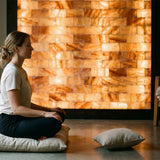 A woman sits cross-legged in profile on a cushion in a dimly lit room, with a glowing stack of natural salt lamps behind her and another cushion on the floor in front.