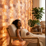 A woman relaxes in a wooden armchair wrapped in a cream knit blanket, with a glowing salt lamp on the table beside her. The background features a warm, illuminated salt wall and potted green plants.