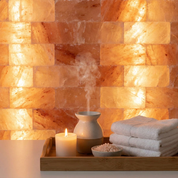 A spa-style scene with a wooden tray holding a lit candle, a small diffuser emitting steam, a bowl of bath salts, and folded white towels in front of a glowing salt lamp wall.