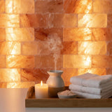 A spa-style scene with a wooden tray holding a lit candle, a small diffuser emitting steam, a bowl of bath salts, and folded white towels in front of a glowing salt lamp wall.