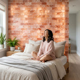 A woman sits cross-legged on a bed in a softly lit bedroom, wearing a light pink knit sweater and shorts. The bed is dressed in cream bedding with a gray throw blanket, and behind it is a large illuminated pink stone wall.