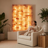 A woman in a beige robe relaxes in a cream armchair, holding a mug, with a large glowing salt lamp wall behind her in a warm, modern room setting.