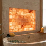A warm-toned salt wall panel made of rectangular illuminated salt blocks mounted above a stone bathtub, with a lit candle on a tray and spa stones and greenery nearby.