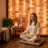 A woman in a cream-colored lounge set sits cross-legged on a woven cushion in a meditative pose, set against a warm, glowing salt wall in a cozy room.