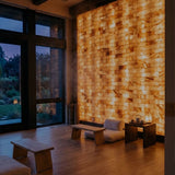 A large illuminated wall made of stacked natural salt blocks glows with warm golden light in a modern interior room with wood floors, low wooden benches, and large windows showing dusk outside.