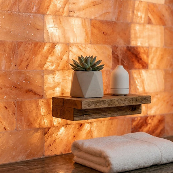 A floating rustic wooden shelf mounted against a glowing warm orange salt wall, holding a small potted succulent in a geometric white planter and a white diffuser. A folded cream towel rests below the shelf.