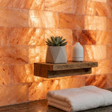 A floating rustic wooden shelf mounted against a glowing warm orange salt wall, holding a small potted succulent in a geometric white planter and a white diffuser. A folded cream towel rests below the shelf.