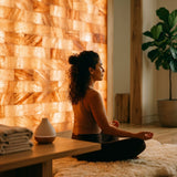 A woman meditates in a serene room with a glowing salt wall, wooden furniture, and a potted plant.
