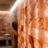 A person standing in a steamy indoor shower or spa room with large illuminated pinkish-orange stone wall panels behind them. A stone bench with folded towels is visible on the left.