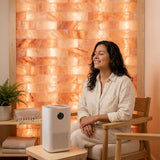A woman sits in a wooden armchair in a spa-like room with a glowing salt wall behind her, with a white air purifier on a small table in the foreground.