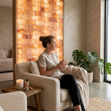 A woman sits in a cream armchair in a spa-like room, wrapped in a beige knit throw and holding a cup, with a glowing orange salt wall behind her, a potted monstera plant to the right, and a small wooden side table with a candle and bottles nearby.