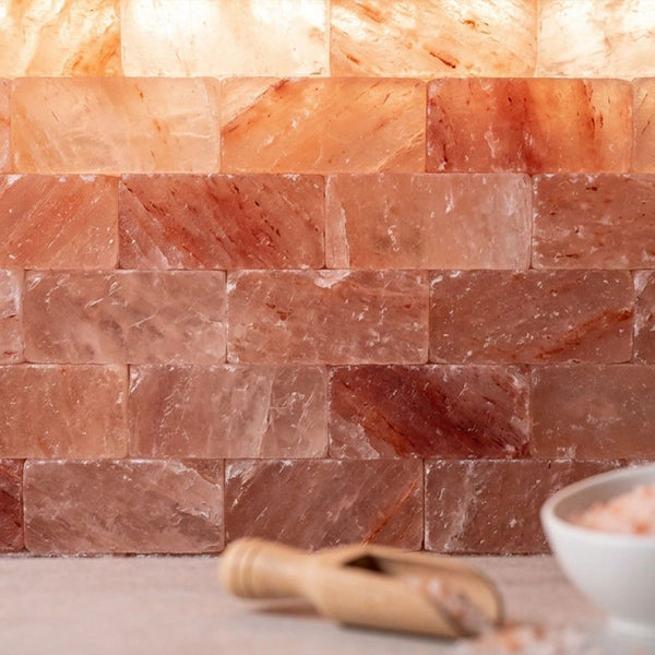 A close-up of stacked pinkish-orange salt blocks arranged like a backsplash, with a small wooden scoop and a white bowl partially visible in the foreground on a light surface.
