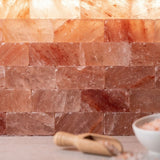 A close-up of stacked pinkish-orange salt blocks arranged like a backsplash, with a small wooden scoop and a white bowl partially visible in the foreground on a light surface.