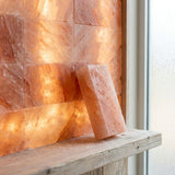 A glowing pink salt lamp made of stacked rectangular salt blocks, with one block leaning against a rustic wooden shelf in front of a bright window.