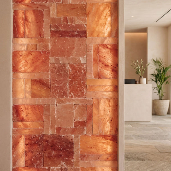 A decorative wall panel made of rectangular polished stone tiles in warm copper, terracotta, and orange-brown tones, shown installed in an interior hallway with a reception desk and potted plants visible in the background.
