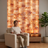 A man sits in a cream armchair in a styled room, with a large illuminated salt lamp wall behind him and a small wooden side table holding glowing crystal lamps and a potted plant.