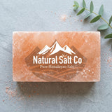 A rectangular bar of Natural Salt Co Pure Himalayan Salt, Brick showing its characteristic pinkish-orange color and white logo, styled on a gray slate background with a green eucalyptus sprig.