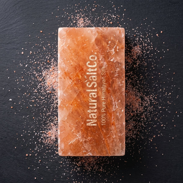 A rectangular bar of Natural Salt Co. Himalayan salt in a warm peach-orange color, standing upright on a dark slate background with scattered salt crystals around it.