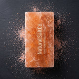 A rectangular bar of Natural Salt Co. Himalayan salt in a warm peach-orange color, standing upright on a dark slate background with scattered salt crystals around it.